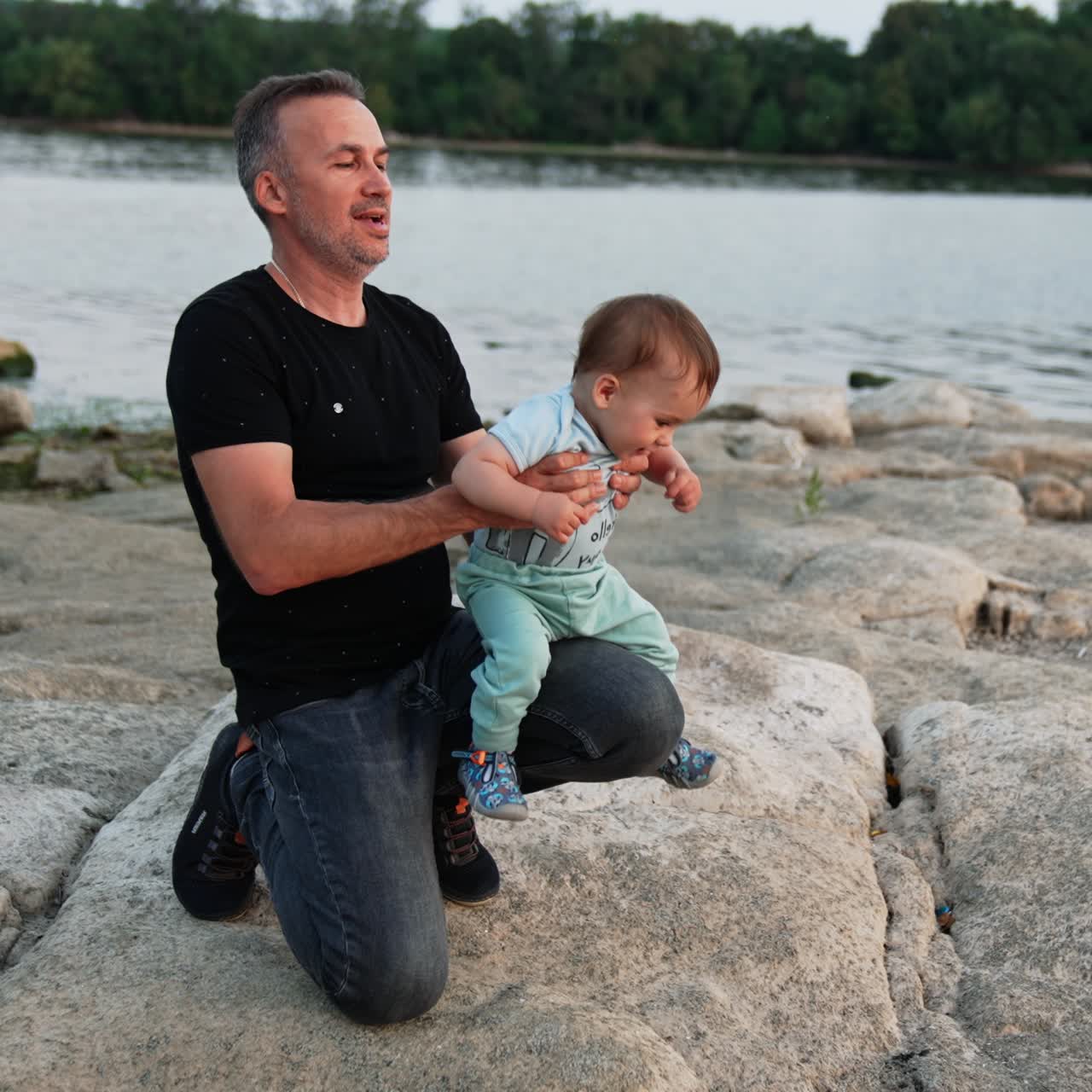 Father holding his little baby boy on one knee. Loving daddy kissing his son's cheek and back of the neck. Family concept. River at backdrop
