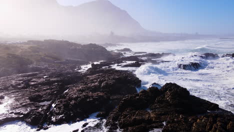 Rocky-coastline-in-Hermanus-pummelled-by-waves,-morning-mist-over-Voëlklip