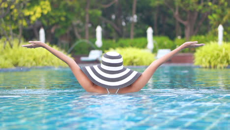 vista trasera de una mujer con sombrero blanco y negro levantando las manos del agua en la piscina sobre un fondo verde borroso a cámara lenta durante el día