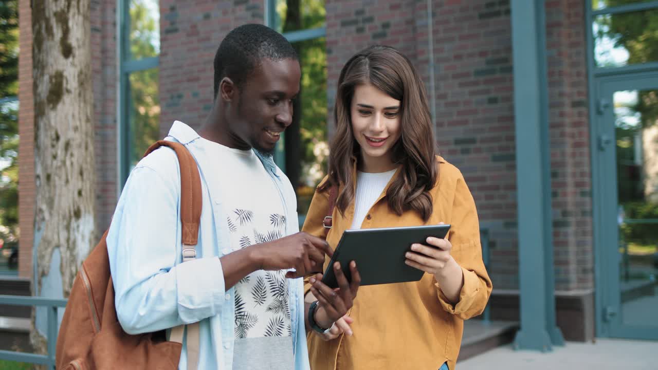mujer caucásica y hombre afroamericano hablando de algo mirando una tableta en la calle cerca de la universidad