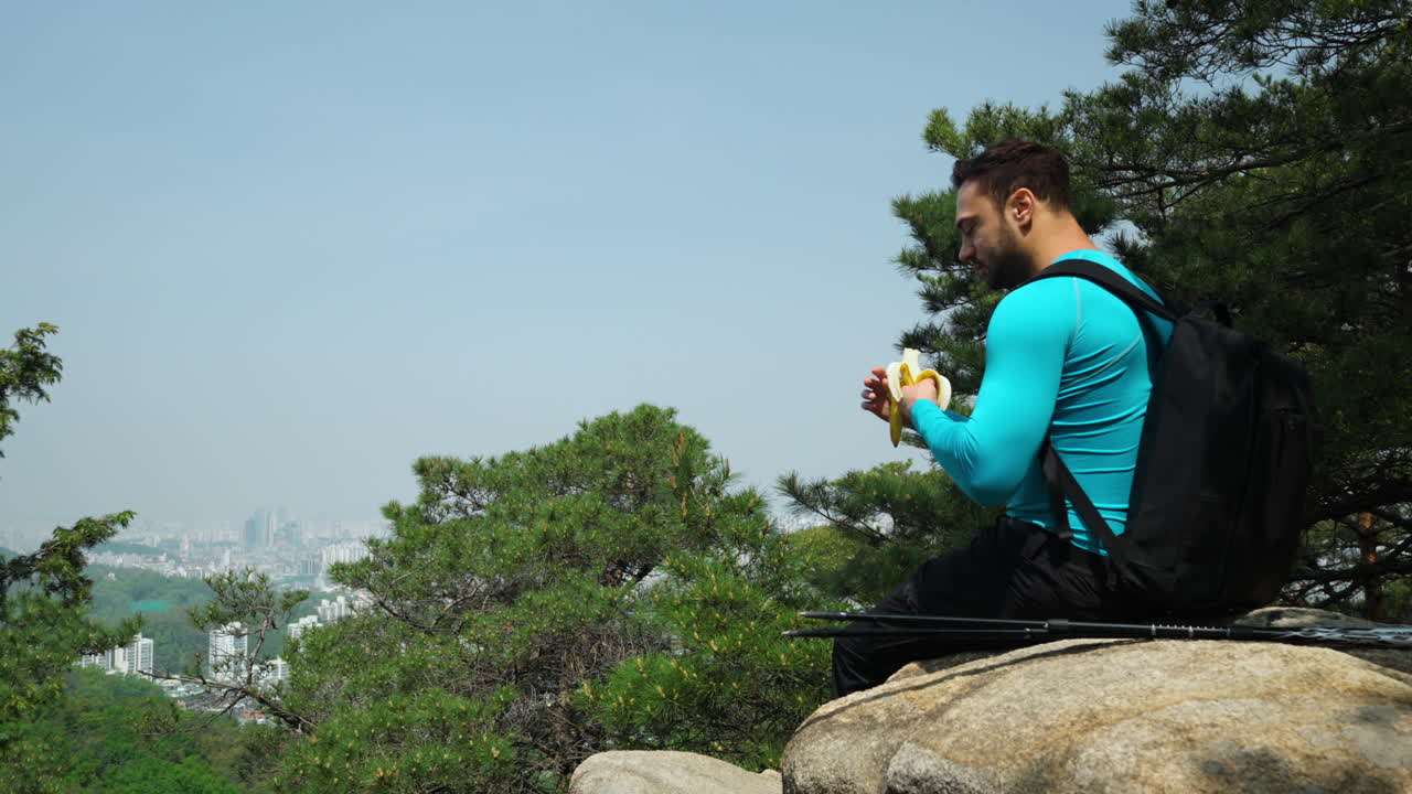 Man Hiking Resting Sitting on Rocky Cliff and Eating Banana Eanjoying Seoul City Skyline from Gwanaksan Mountain