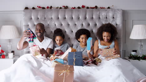 Joven-Familia-De-Raza-Mixta-Sentados-Juntos-En-La-Cama-Desenvolviendo-Regalos-En-La-Mañana-De-Navidad,-Vista-Frontal