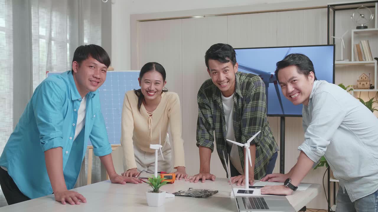 아시아 엔지니어 그룹이 사무실에서 풍력 터빈에 대해 작업하는 동안 카메라에 미소 짓고 있습니다.