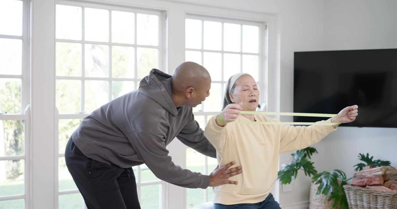 African American coach approaching guiding senior Asian stretching band on ball at home for balance