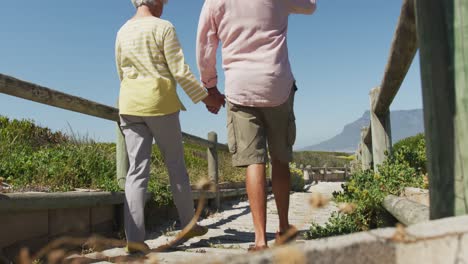 Rückansicht-Eines-älteren-Kaukasischen-Paares,-Das-Händchen-Hält-Und-Auf-Dem-Weg-Zum-Strand-Geht