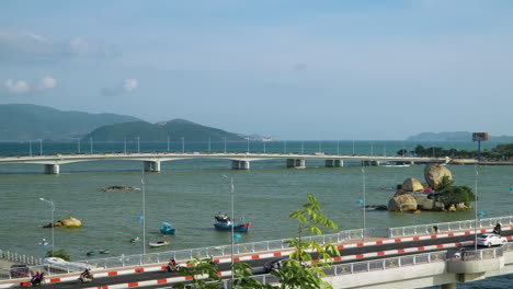 vehicle traffic on tran phu and xom bong bridges over cai river in nha trang on sunny day, wooden fishing boats anchor on water at cu lao village, vietnam