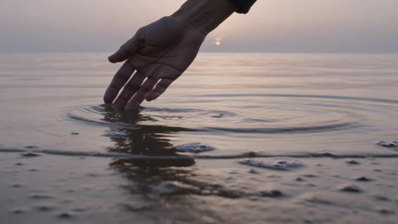 Mano e avambraccio che sfiorano l'acqua bassa a riva, con una manica scura che crea increspature, entrando nell'inquadratura.