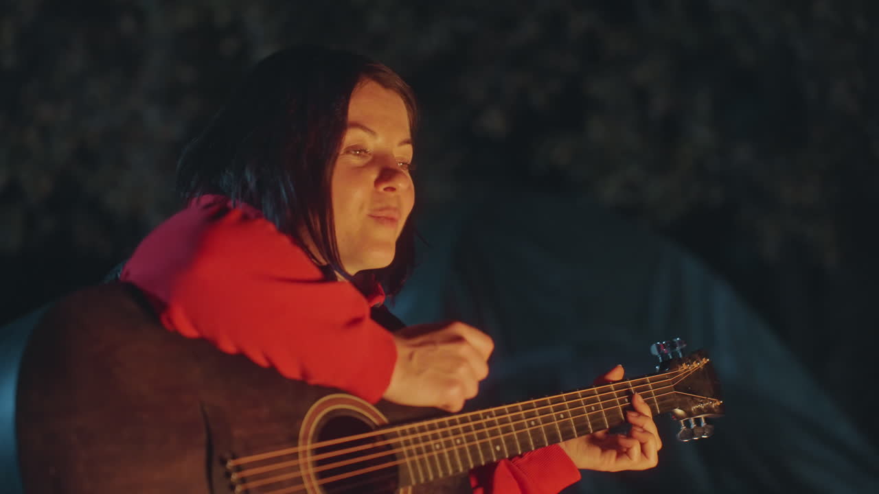 Cantante intimo con melodia da falò. Scena serena di un musicista che suona la chitarra accanto a un falò tremolante. Atmosfera serale calma con un musicista che strimpella la chitarra vicino a una tenda sotto la calda luce del fuoco