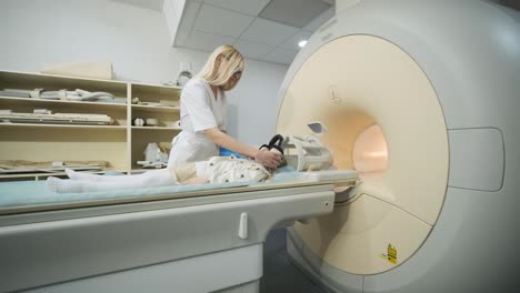 doctor radiologist puts noise isolating headphones on the patient little girl to make an mri scanning of brain, head, neck, girl lies on automatic table, using modern equipment, coil on the head