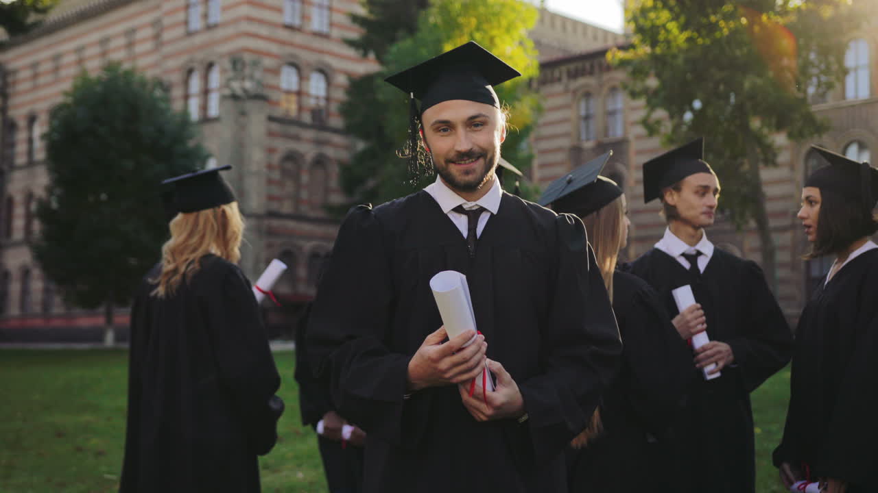 Portrait shot of a young man in graduation gown and cap looking at the diloma in his hands and then to the camera