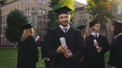 Portrait-shot-of-a-young-man-in-graduation-gown-and-cap-looking-at-the-diloma-in-his-hands-and-then-to-the-camera