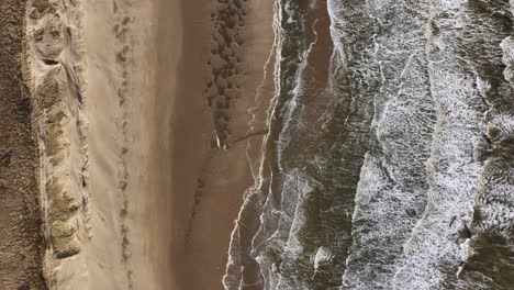 Experience-the-raw-power-of-the-ocean-as-waves-fiercely-crash-onto-a-sandy-beach,-surrounded-by-rolling-dunes-and-a-backdrop-of-brown-bushes-in-this-stunning-aerial-footage