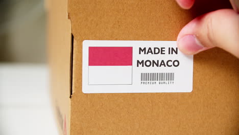 Hands-applying-MADE-IN-MONOCO-flag-label-on-a-shipping-cardboard-box-with-products