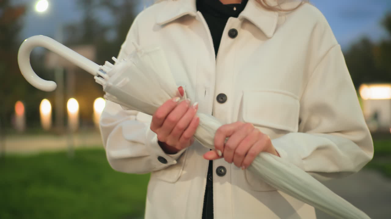 Close up of woman in white coat holding folded umbrella with both hands while standing outdoors during evening with soft blurred city lights glowing in background