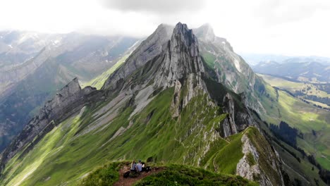 Aerial-flyover-over-the-two-hikers-enjoying-the-view-of-Schafler-ridge-in-Appenzell,-Switzerland-with-cliffs-and-mountain-peaks-in-view