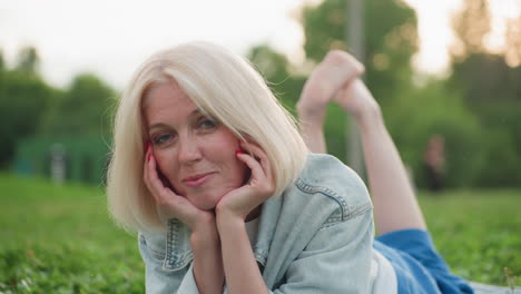 stylish woman lying on green grass in park resting chin on hands, thoughtful gaze, casual denim jacket, soft background of blurred trees and warm light, relaxed mood conveying serenity