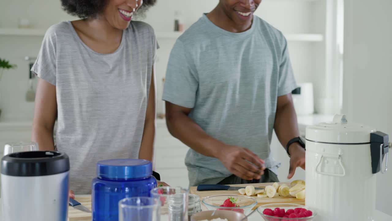Lächelnd beginnt ein Gespräch, zwei Partner schneiden Bananen und Beeren und geben sie für einen Smoothie in den Mixer.