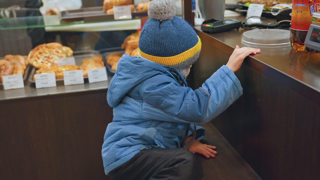 Figlio caucasico che si protende verso la vetrina della panetteria con la mamma nelle vicinanze, un bambino curioso con cappotto blu e berretto a righe gialle che si allunga per afferrare il bordo del bancone e sbirciare i dolci, la madre sta vicina e attenta.