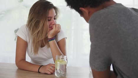 joven mujer caucásica con un vaso de agua sentada junto a la mesa hablando con su novio africano. familia feliz disfrutar