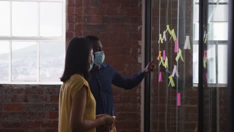 diverse male and female business colleagues wearing face masks brainstorming pointing to glass wall