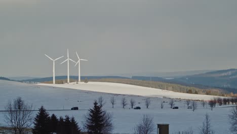 Three-windmills-in-snow-white-landscape,-cars-passing-on-road,-Klinovec,-Czech-Republic