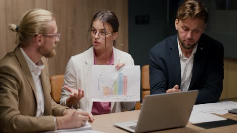 A-brunette-girl-in-a-white-suit-in-a-pink-shirt-and-round-glasses-shows-a-graph-on-a-sheet-of-paper-to-her-interlocutor-on-a