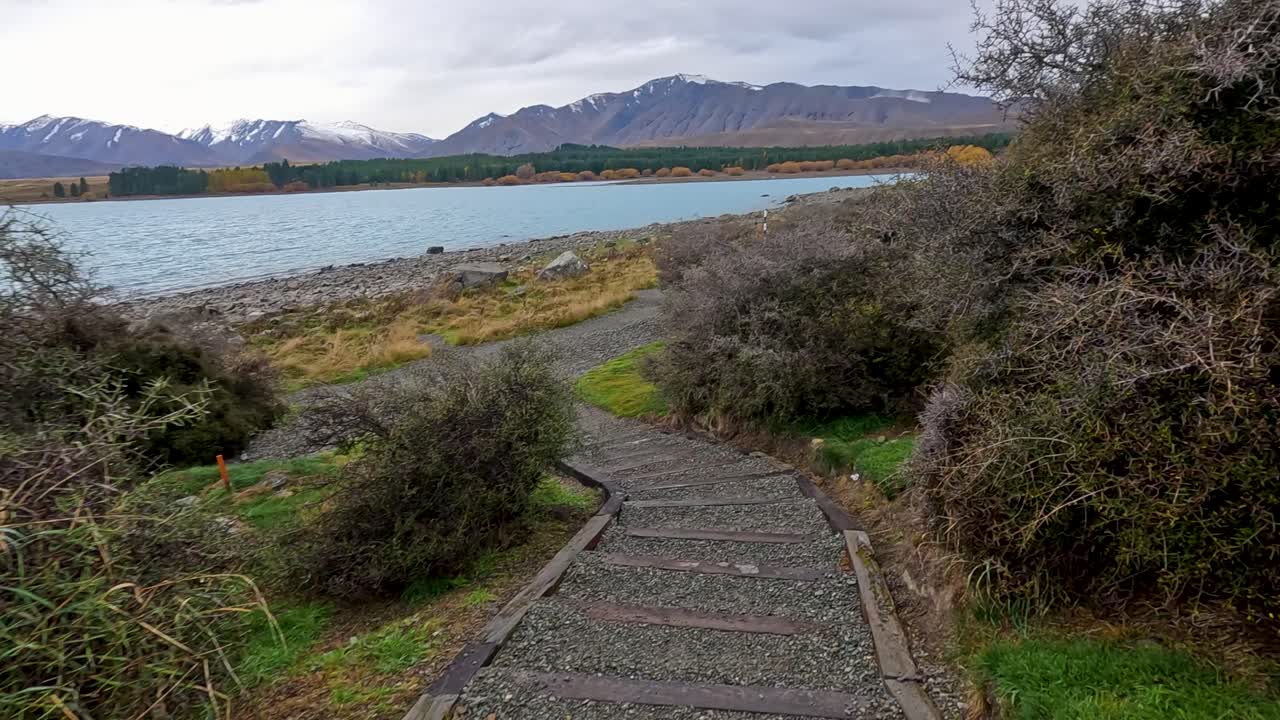 A steady camera moves down a gravel stairway path lined with shrubs, revealing a tranquil lake and distant mountains under overcast daylight