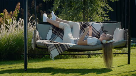 a teenage girl with long hair listens to music on headphones, lies on a garden swing in the backyard of a house.