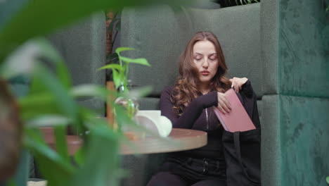 blonde woman dressed in black outfit sits comfortably in green booth reaching into black bag to retrieve pink books while surrounded by indoor plants and soft natural lighting in serene modern setting
