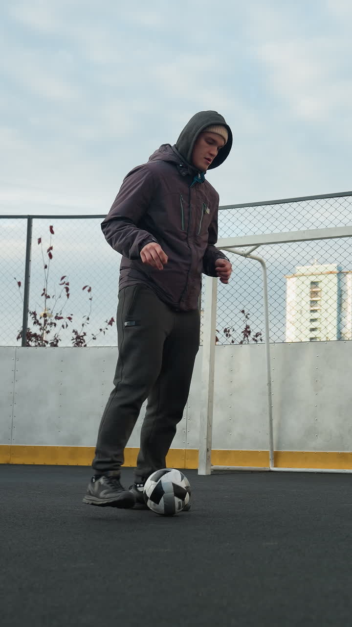 Man practicing soccer drills on sport arena, skillfully controlling ball while focusing on footwork, background includes urban residential buildings, mesh fencing, and goalpost under soft daylight