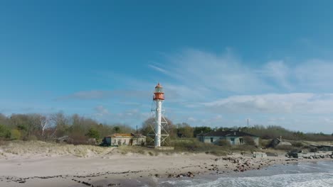 aerial establishing view of white colored pape lighthouse, baltic sea coastline, latvia, white sand beach, large waves crashing, sunny day with clouds, low drone orbit shot