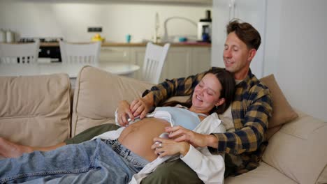 Joyful-man-and-woman-putting-wireless-headphones-on-pregnant-woman-tummy-while-relaxing-at-home