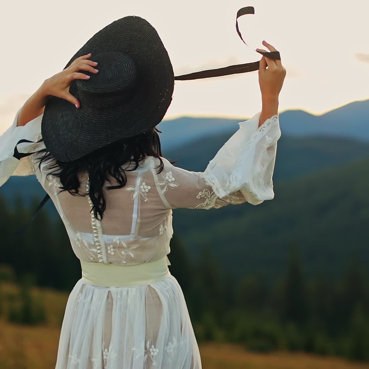 Young lady in white dress and black hat stands her back to the camera facing setting sun. Woman holding her hat string fluttering in the wind. Mountains in blur at backdrop