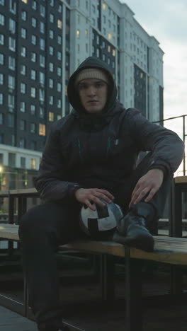 hombre con capucha sentado en un banco girando una pelota de fútbol con la mano izquierda en la pierna durante la sesión de entrenamiento, rodeado de edificios residenciales modernos que brillan con luces de la noche