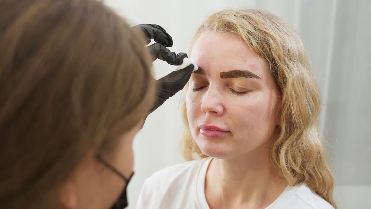 Beautician wearing black gloves uses cotton pad to clean eyebrow area of client with blond hair during beauty treatment session, both focused in clean bright salon environment with gentle care