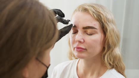 beautician wearing black gloves uses cotton pad to clean eyebrow area of client with blond hair during beauty treatment session, both focused in clean bright salon environment with gentle care