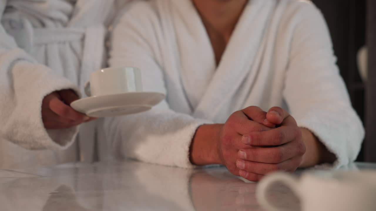 Woman serves man with coffee in kitchen closeup. Young girlfriend in bathrobe gives cup of hot drink to beloved boyfriend at home in morning