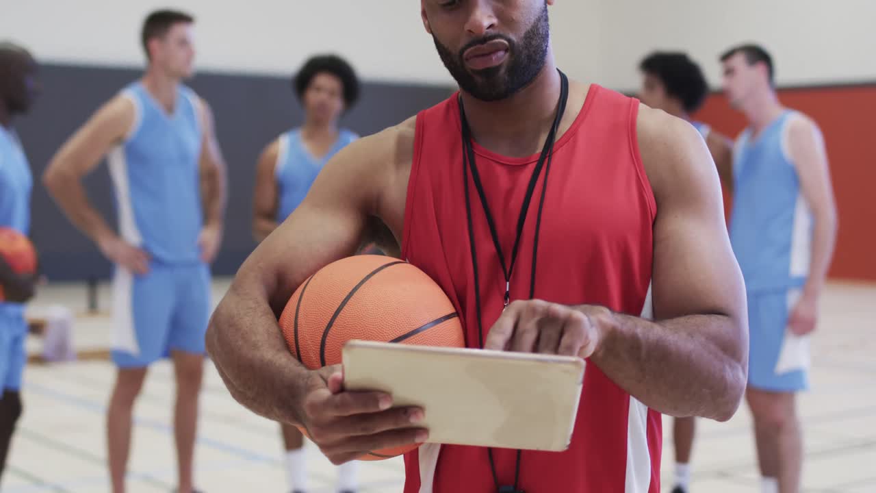 entrenador de baloncesto masculino biracial usando una tableta en una cancha cubierta con un equipo masculino diverso, en cámara lenta