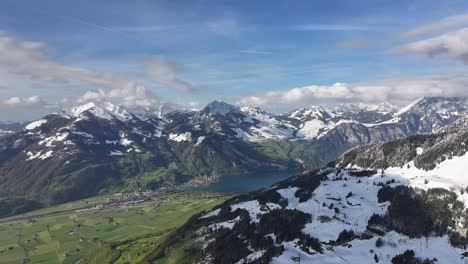 slow aerial dolly towards snow covered mountain peaks with village and blue lake in foreground