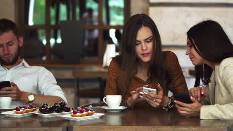 Reunión-Uno-A-Uno.-Dos-Jóvenes-Mujeres-De-Negocios-Sentadas-A-La-Mesa-En-Un-Café.-La-Chica-Muestra-La-Imagen-De-Su-Amiga-En-La-Pantalla-Del-Teléfono-Inteligente.-Sobre-La-Mesa-Hay-Un-Cuaderno-Cerrado.-Reunión-Con-Amigos.