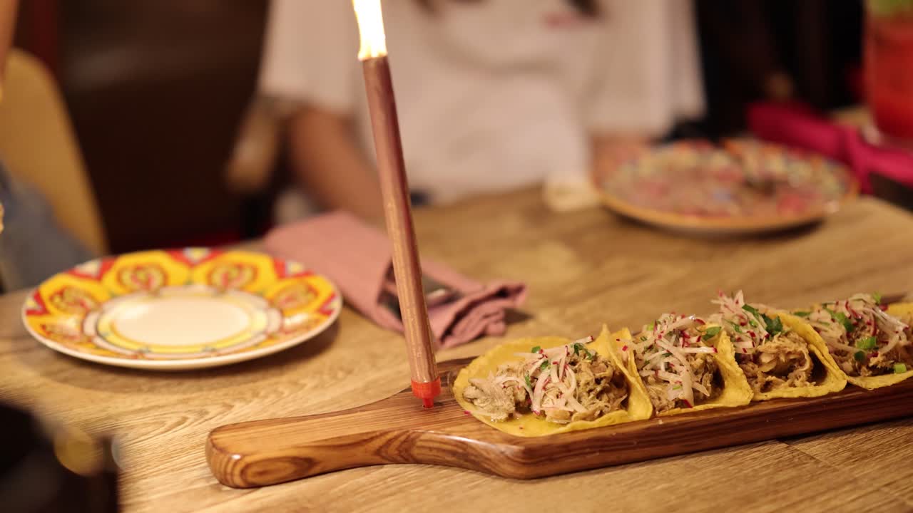 A wooden platter of assorted tacos on a warmly lit table, surrounded by plates and drinks in a restaurant