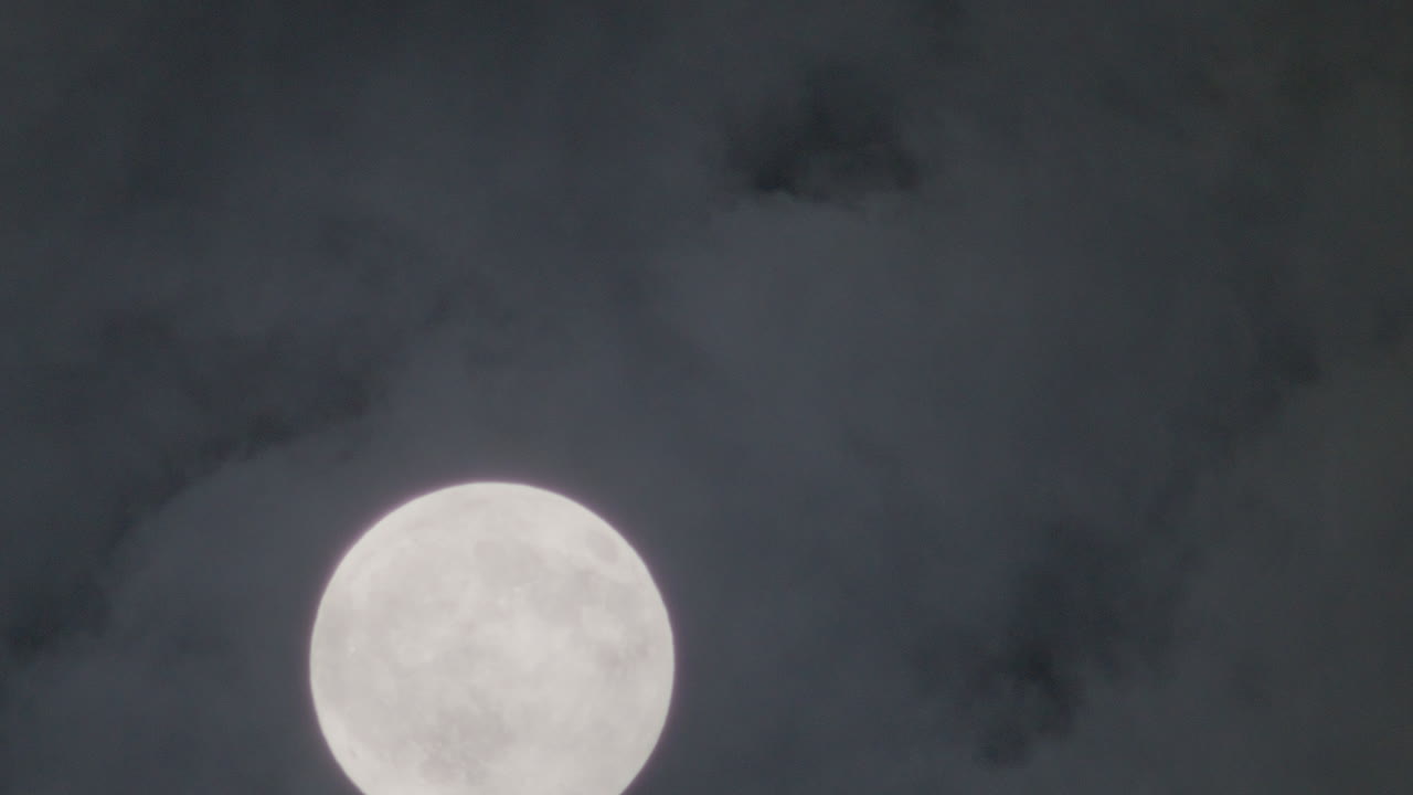 The full moon slowly ascends through dark clouds in a moody night sky, marking the quiet onset of nighttime