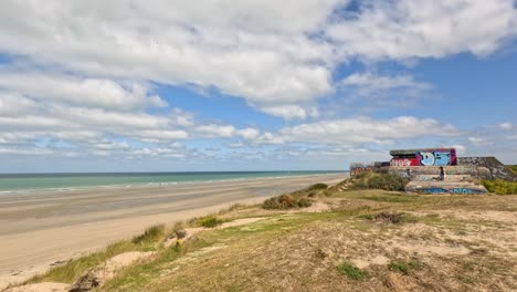 A wide daylight pan across a historic coastal fortification with graffiti, sandy grassland, and sea under partly cloudy skies near Dunkirk, France