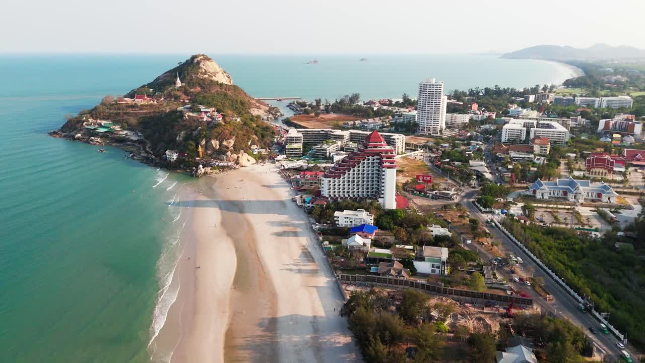 Drone aerial overview of tropical Hua Hin coastline destination, beachfront resorts, turquoise waters, and city skyline on the hazy background