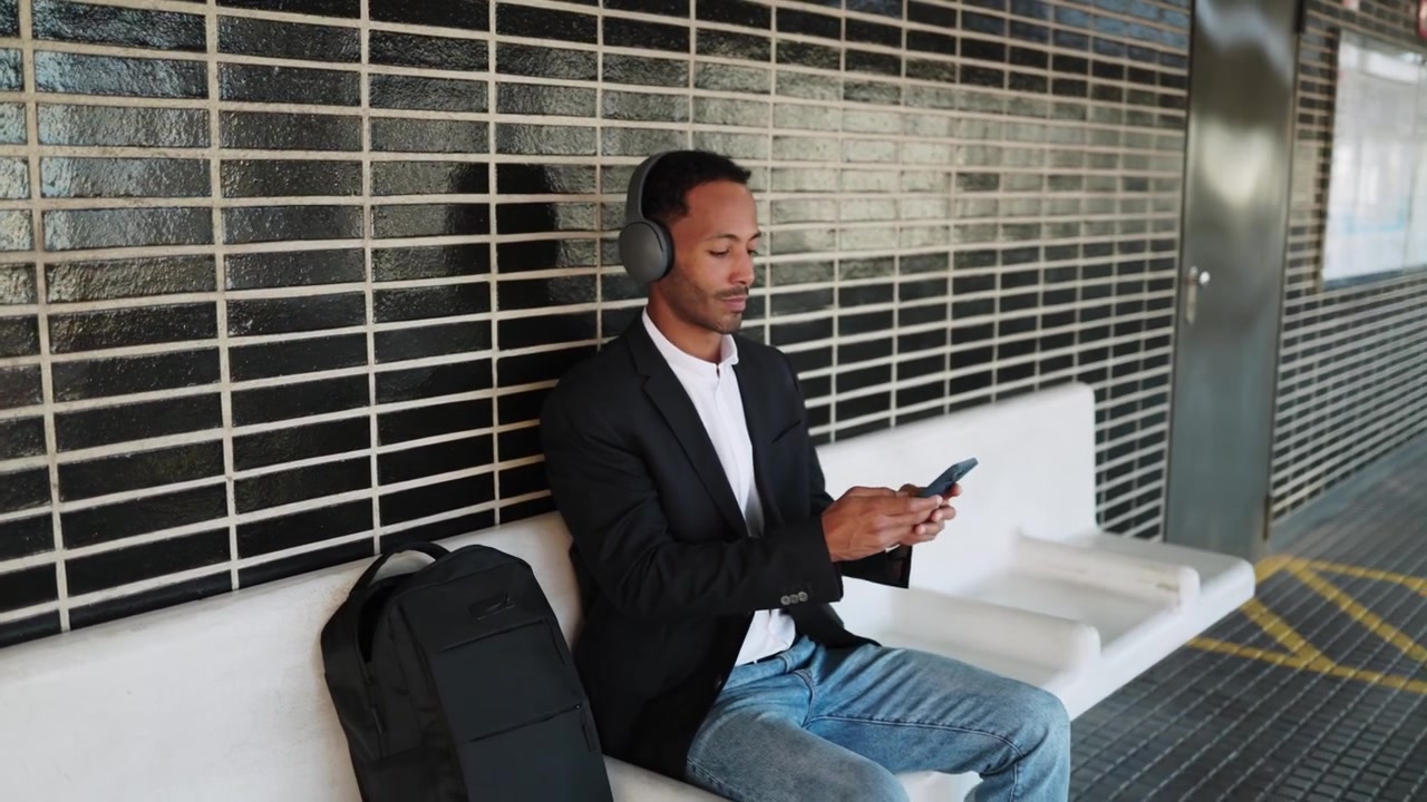 Young Man Sitting on Bench at Train Station Using Smartphone