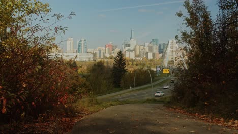 Una-Tranquila-Vista-Del-Horizonte-De-Edmonton,-Alberta-En-El-Otoño-Mientras-Los-Autos-Ingresan-Al-Centro-De-La-Ciudad,-Todo-Visto-A-Través-De-Un-Tranquilo-Sendero-Del-Parque-Donde-Los-árboles-Rojos-Y-Amarillos-Comienzan-A-Caer-De-Las-Ramas