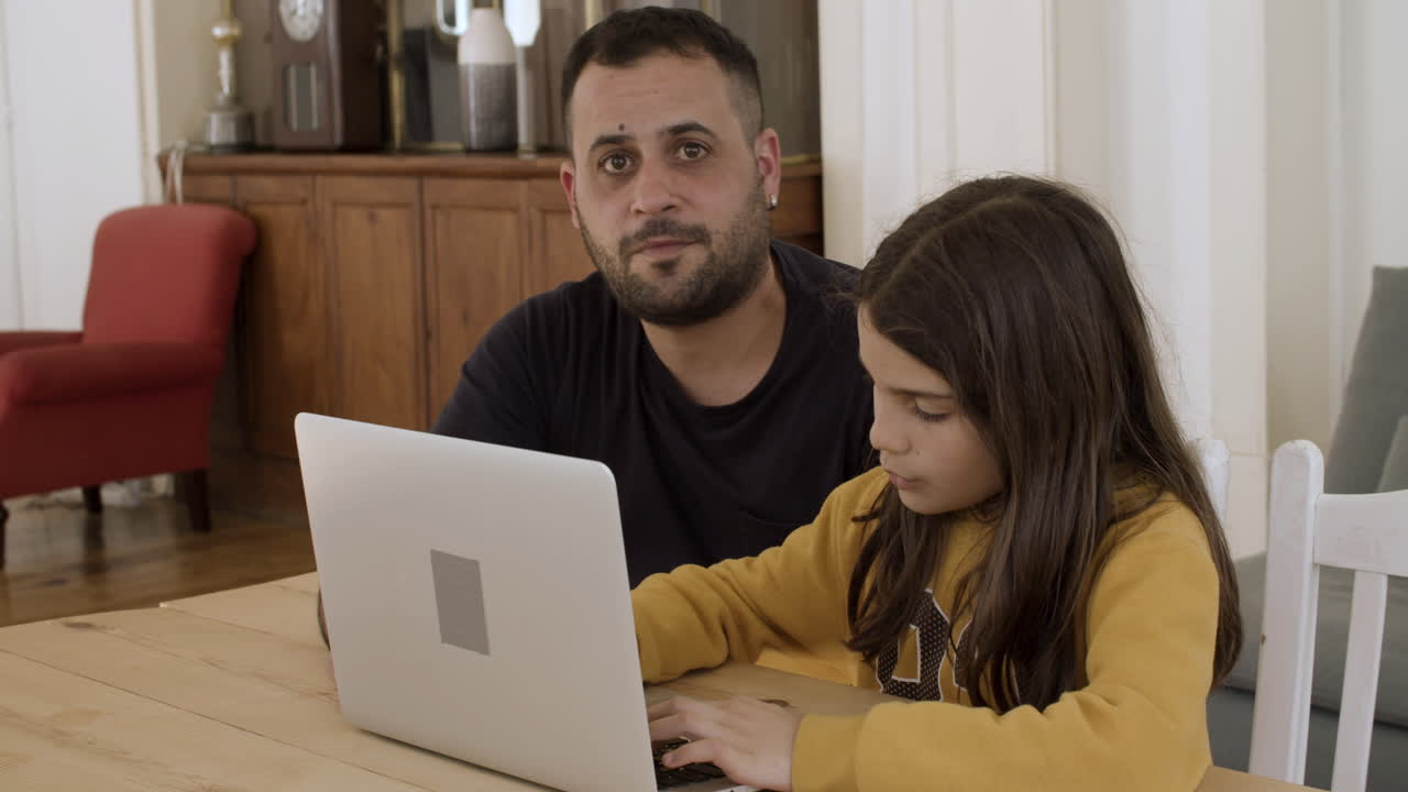 padre y hija caucásicos sentados en la mesa, usando la computadora portátil.
