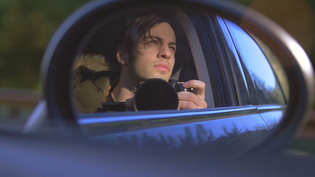 Man in Vehicle Sideview Mirror. A young man looks into the mirror of a driving car. The sun shines on his face, the terrain passes by. Captured in slow motion.
