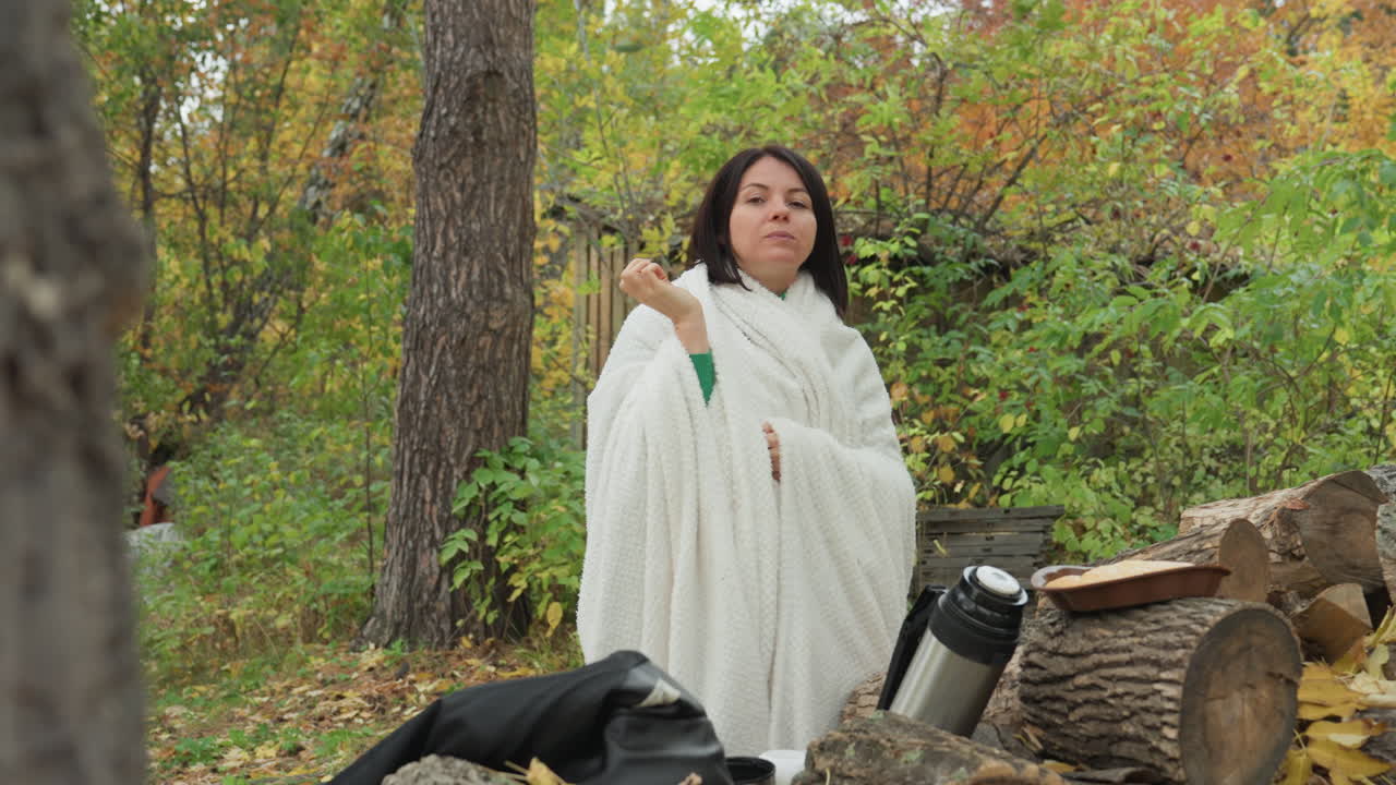 Donna avvolta in una coperta nella foresta che sorseggia da un thermos appoggiata a un tronco vicino a una giacca di pelle e legna da ardere, fogliame autunnale, atmosfera tranquilla e contemplativa, trama a maglia confortevole, scena di rifugio rustico