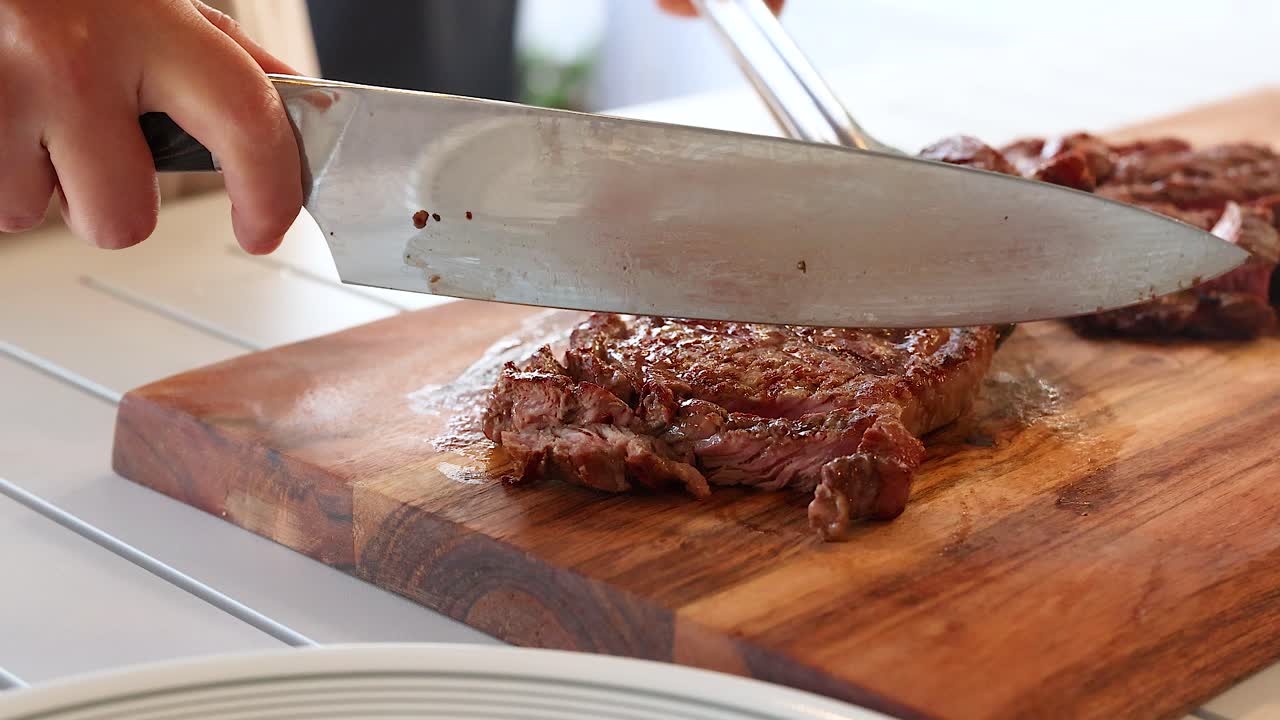 Chef slices cooked beef steak on a wooden board using a large knife and fork. Bright kitchen setting